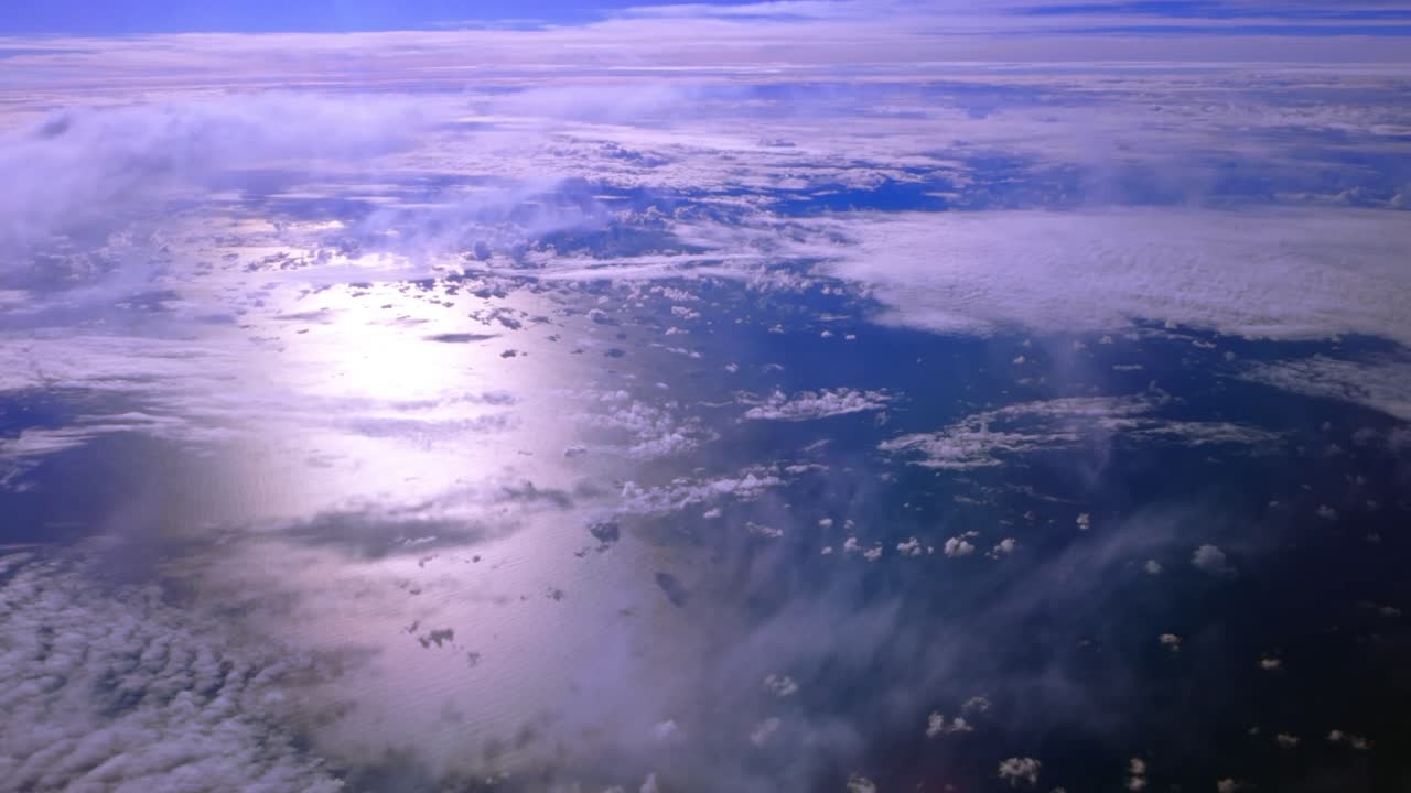 A high altitude aerial view over clouds and water on a sunny day, taken from the cabin of a passenger aircraft. The camera is stationary looking down out the port side window at the ocean and clouds.