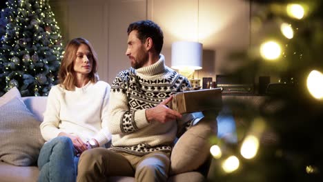 Cheerful-Surprised-Wife-Receiving-Xmas-Present-From-Loving-Caring-Husband-At-Home-On-Christmas-Eve-Near-Decorated-Tree-With-Twinkle-Lights