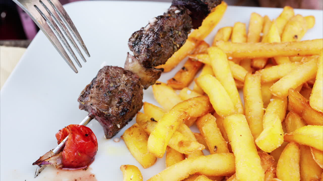 Close up of a woman taking grilled meat off skewers on a white plate with french fries at a restaurant