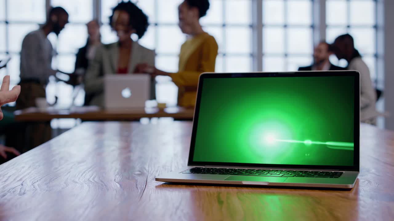 Close-up of a laptop on a wooden table with a green screen, blurred group in background