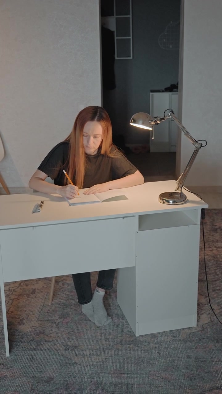 Young woman with long hair writes in open notebook. Woman wears dark clothing and light colored socks maintaining. Setting includes patterned carpeting minimal background details