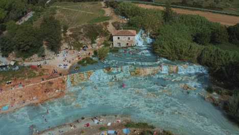 Drohnenaufnahmen-Aus-Der-Luft-Der-Berühmten-Thermalquellen-Von-Saturnia-Cascate-Del-Mulino-Mit-Ihrem-Blauen,-Warmen-Wasser-In-Der-Idyllischen-Landschaft-Der-Toskana,-Italien,-Mit-Hügeln,-Feldern,-Menschen-Und-Olivenbäumen