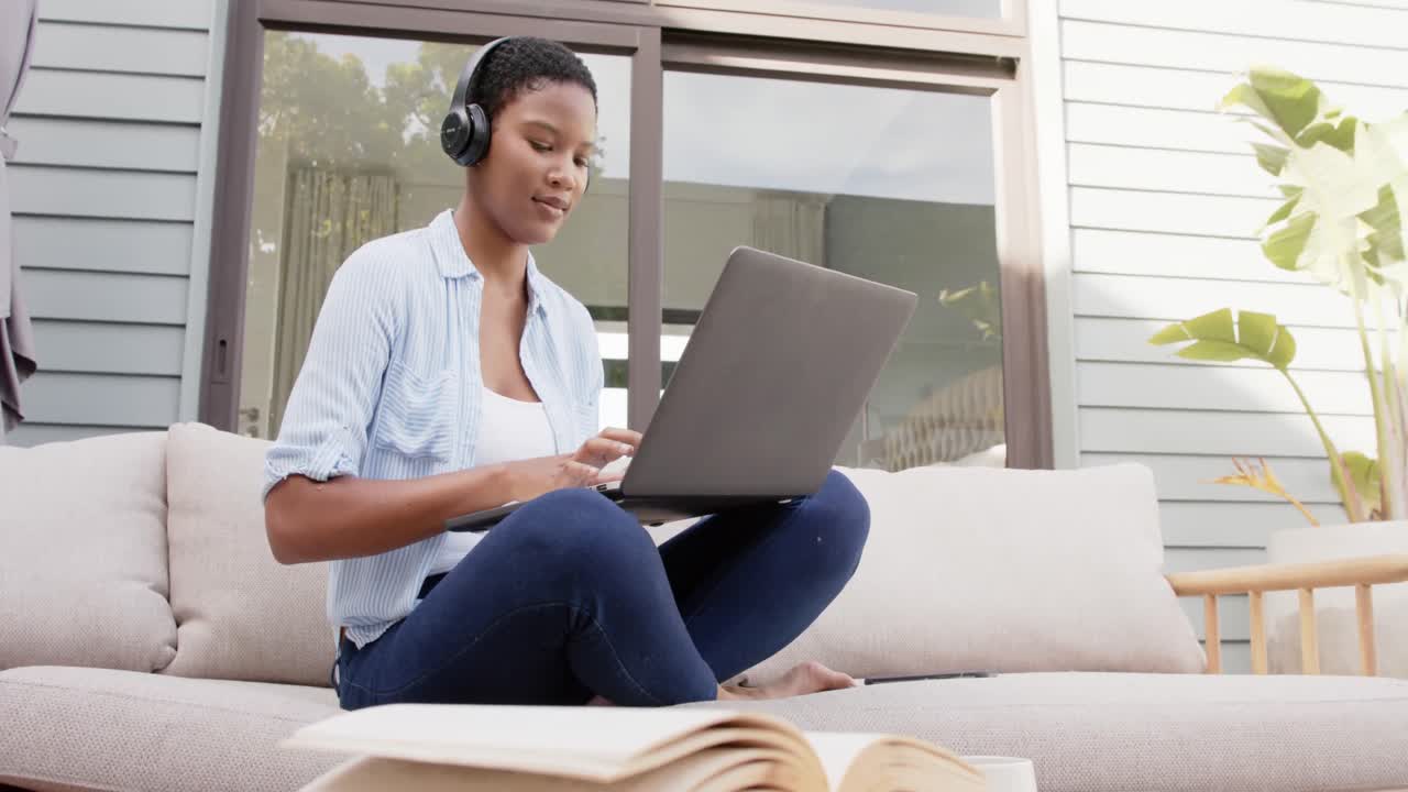 African american woman sitting on sofa on terrace wearing headphones, using laptop, slow motion