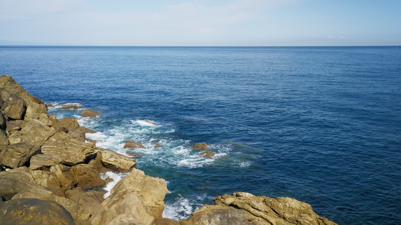 Wide slow motion pan of rough, layered coastal rocks meeting the deep blue Atlantic ocean horizon