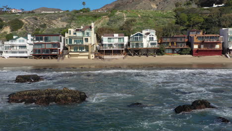 Aerial-shot-of-ocean-waves-and-cliff---waves-washing-up-on-a-rock-in-sea-and-houses-in-the-background