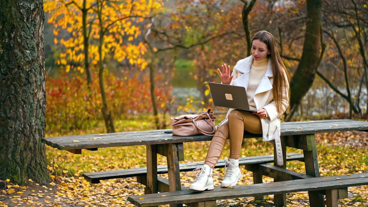 Long-haired brunette lady sitting in the park on wooden table using her laptop. Smiling woman greeting someone in online video chat. Autumn nature backdrop.