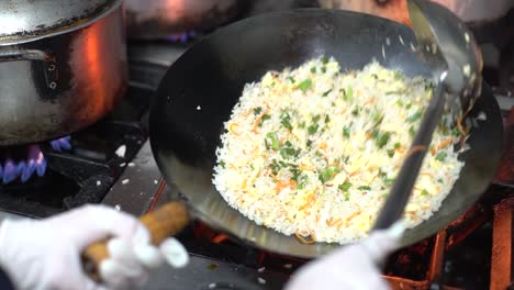 chef in black uniform and sterile gloves, adding spring onions on chao fan or chinese fried rice then mixing, stirring, and tossing it using a wok and a ladle to stir on a full blast fire in a kitchen