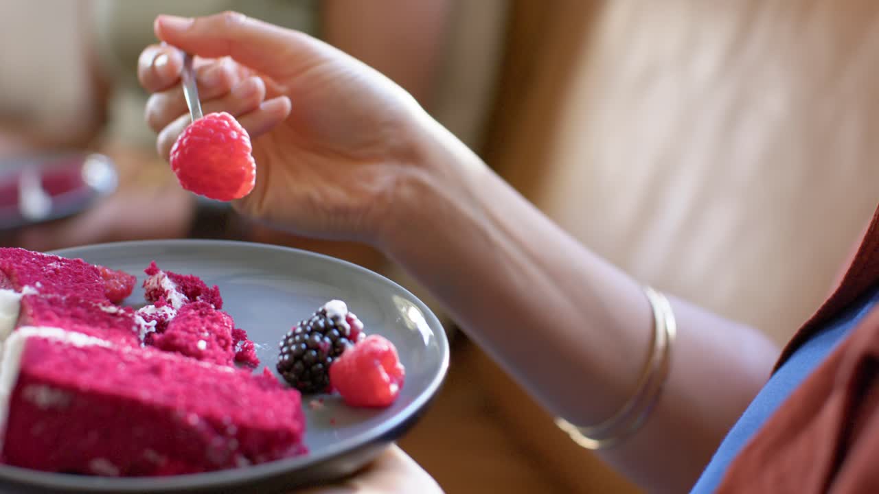 Woman is spearing raspberry on magenta cake plate on lap as fork is settling, to eat