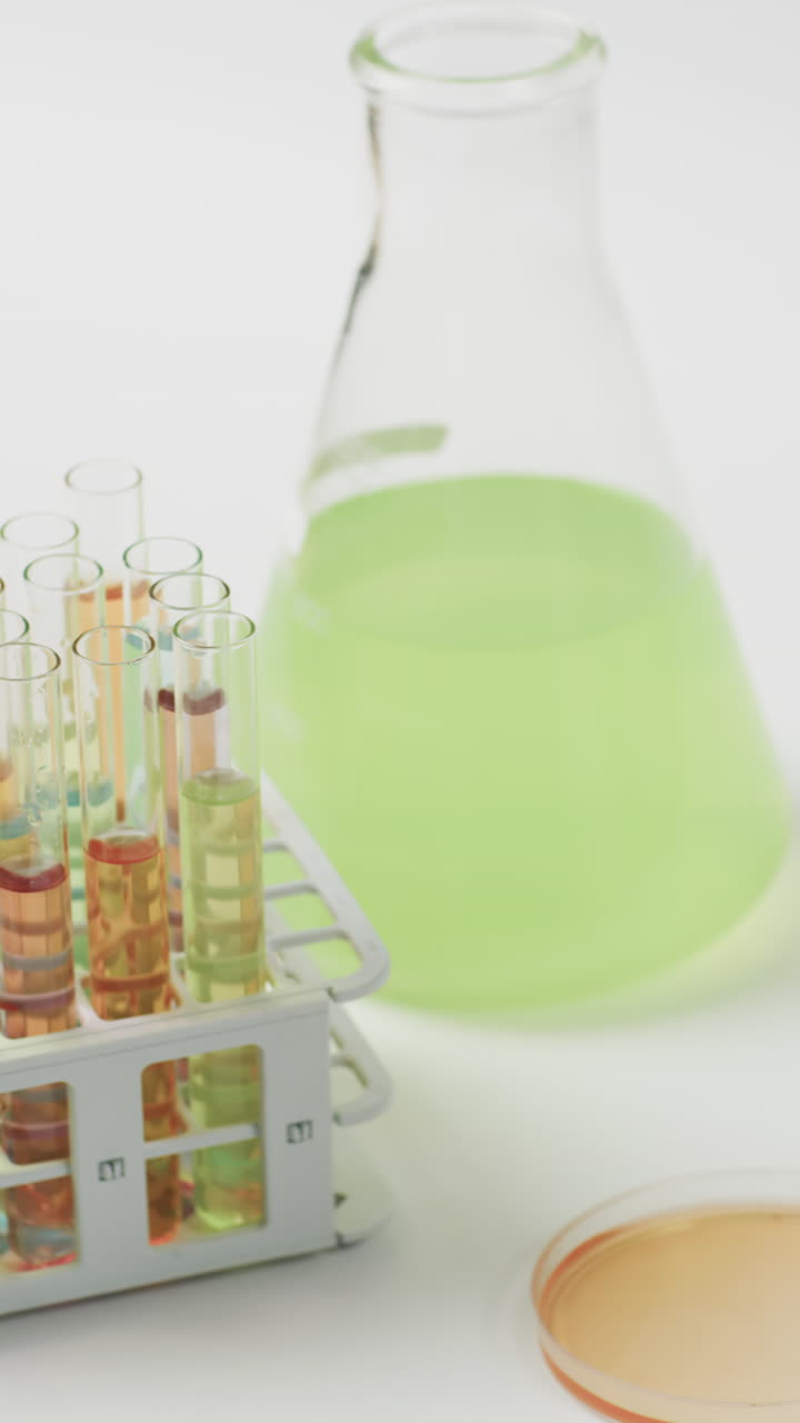 Vertical video of coloured liquid in flask with test tubes in stand on white background