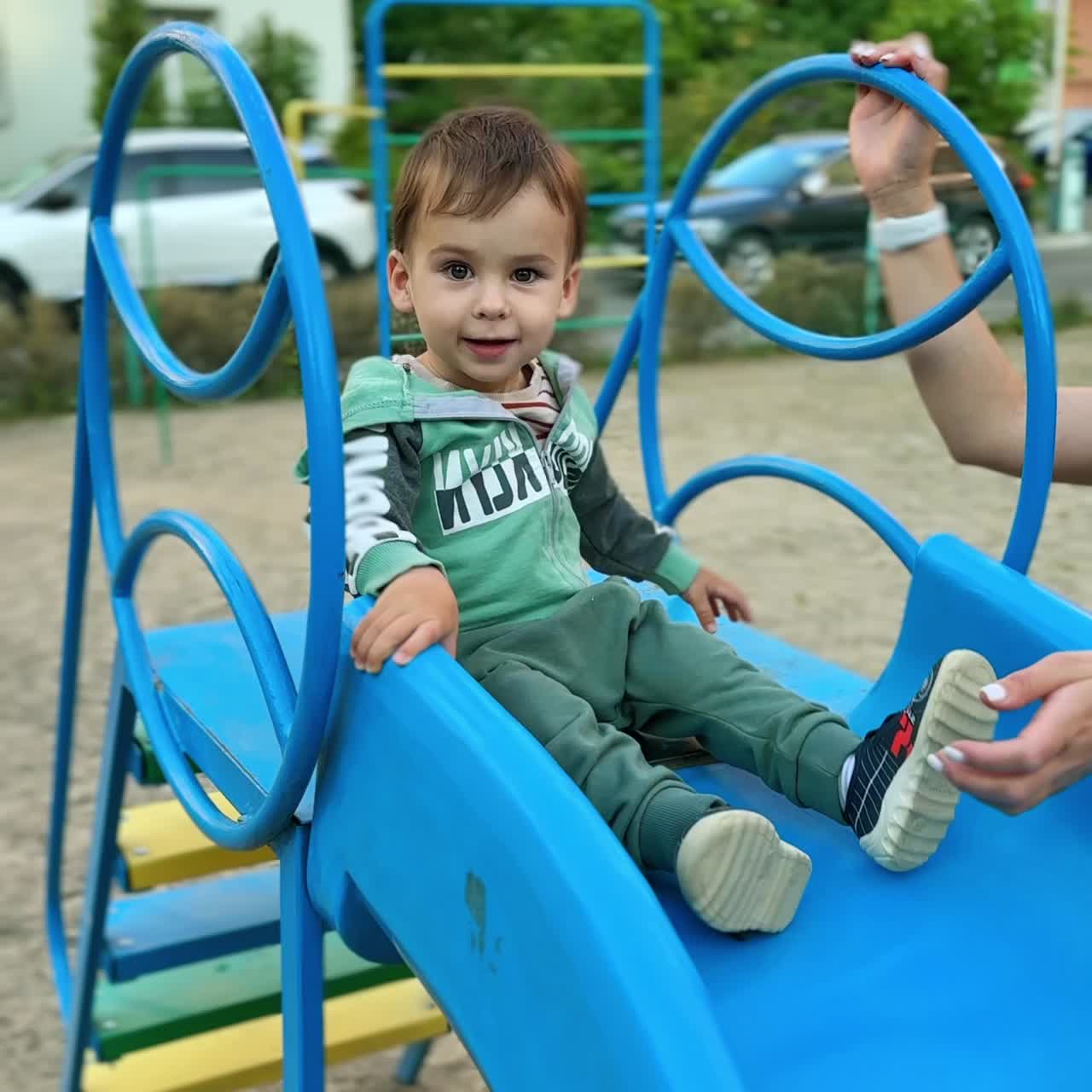 Active energetic Caucasian baby boy sits on the slide waving feet. Kid slides down supported by mom. Vertical screen