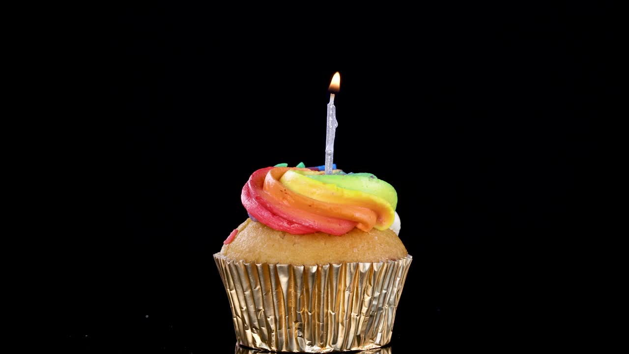 Cupcake with rainbow frosting and lit candle, centered on black background under soft studio lighting