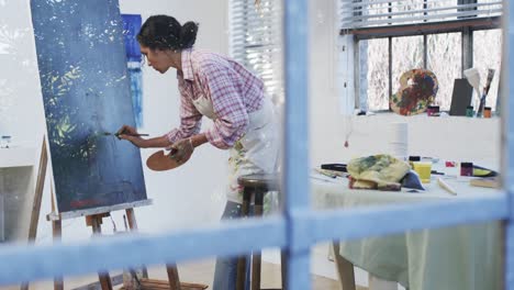 Focused-biracial-female-artist-in-apron-painting-on-canvas-in-studio,-slow-motion