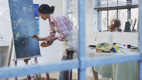 focused biracial female artist in apron painting on canvas in studio, slow motion