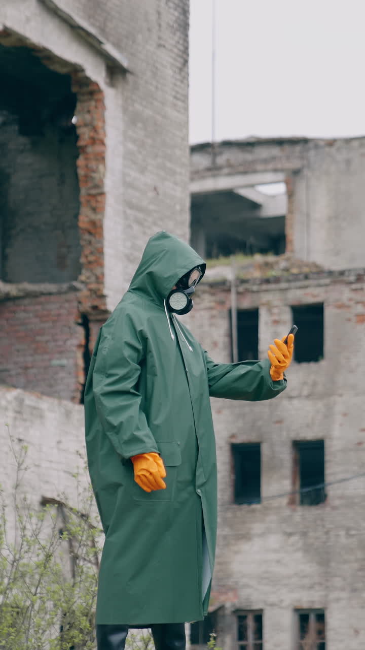 Human in protective suit and gas mask in abandoned place. Survivor holding a mobile phone while standing in ruined city. Post-apocalyptic world after a nuclear war. Vertical video