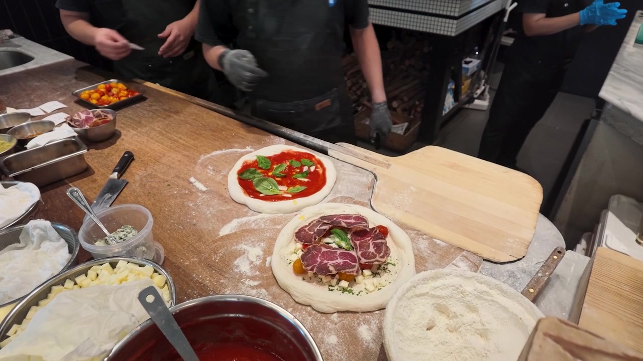 Aufsicht auf Koch mit schwarzer Schürze, der Pizzen mit frischen Zutaten in der Küche zubereitet.