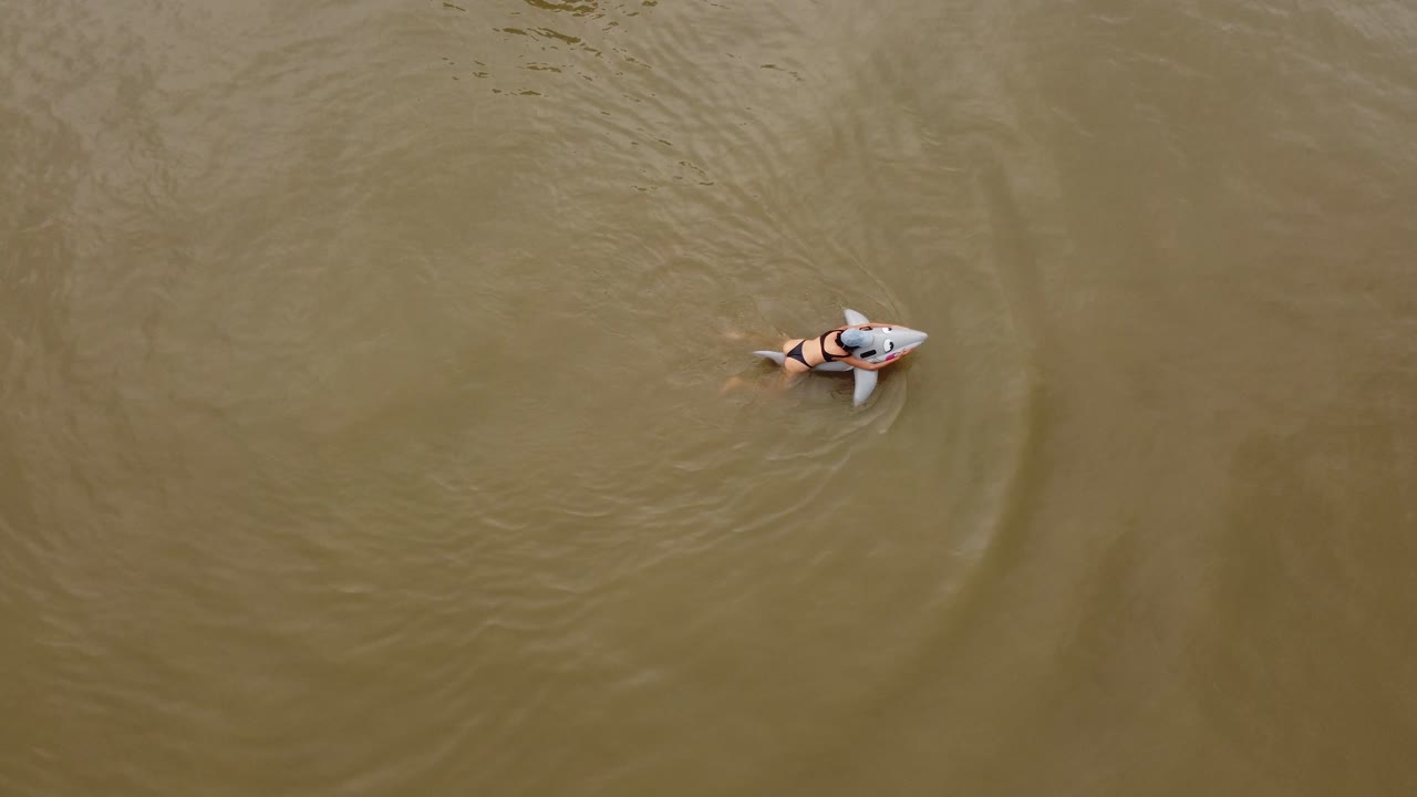 una toma de órbita dinámica de una mujer nadando en un bikini montando en un juguete de agua de tiburón inflable