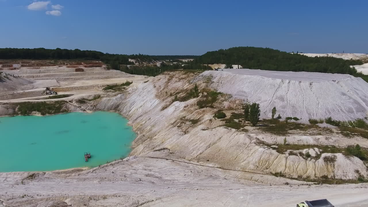 Hilly and sloping area of a kaolin quarry producing raw kaolin. Little blue lake reflecting the sky. Green forest backdrop.