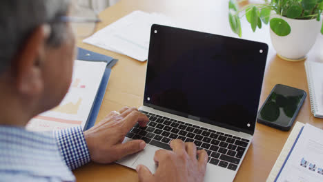 Senior-biracial-male-doctor-sitting-at-desk-using-laptop,-copy-space-on-screen