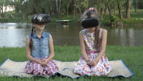 two little girls play and explore the surrounding space with virtual reality glasses. they sit in the park on the grass, against the background of the river and green trees, on a summer evening. 4k.