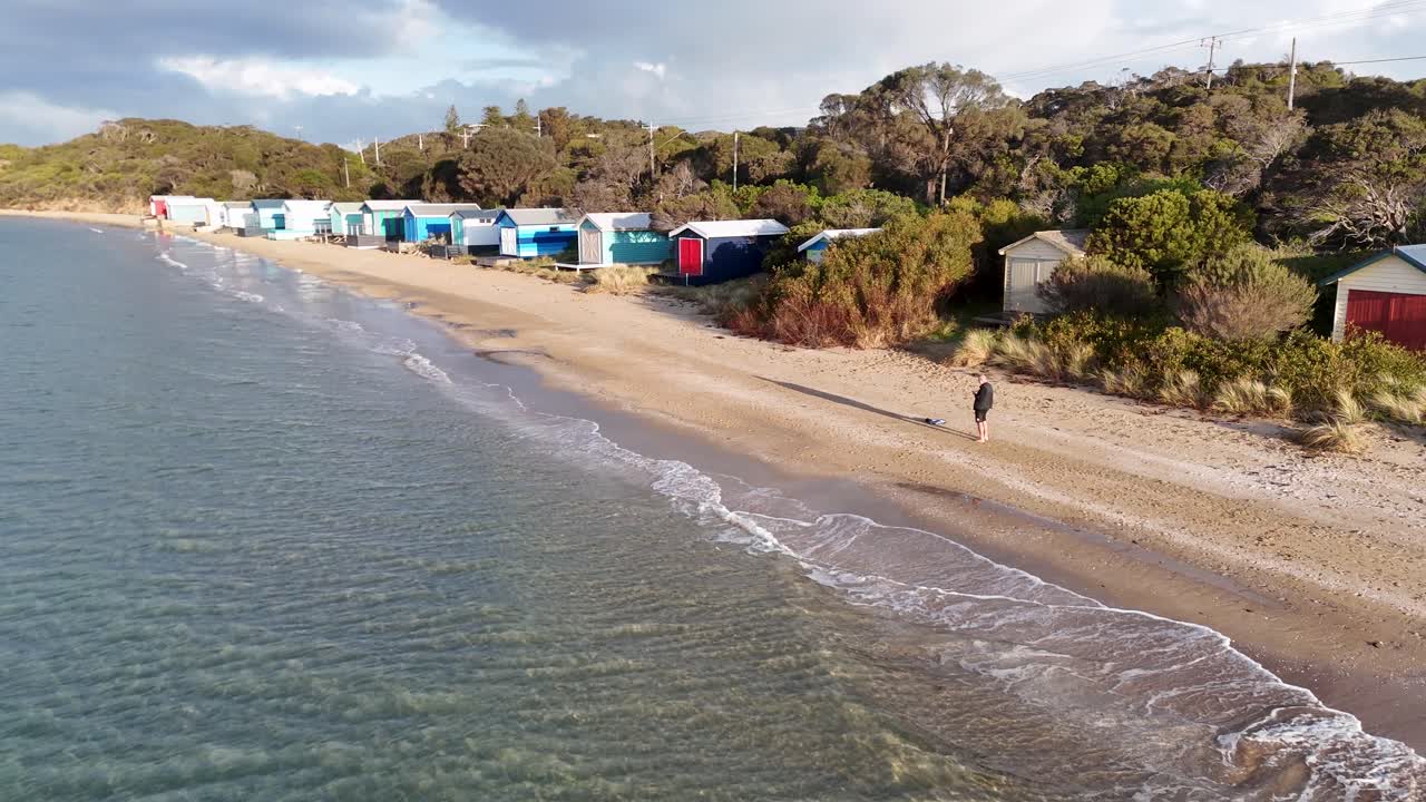 Drone glides above sandy beach with colorful bathing boxes, gentle waves, and soft afternoon sunlight