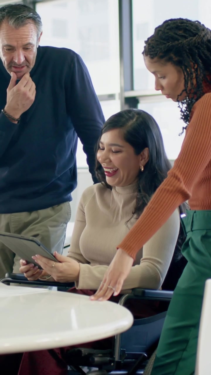 Video verticale: Donna seduta con un top beige che mostra un tablet a un tavolo rotondo vicino alle finestre, condividendo idee.