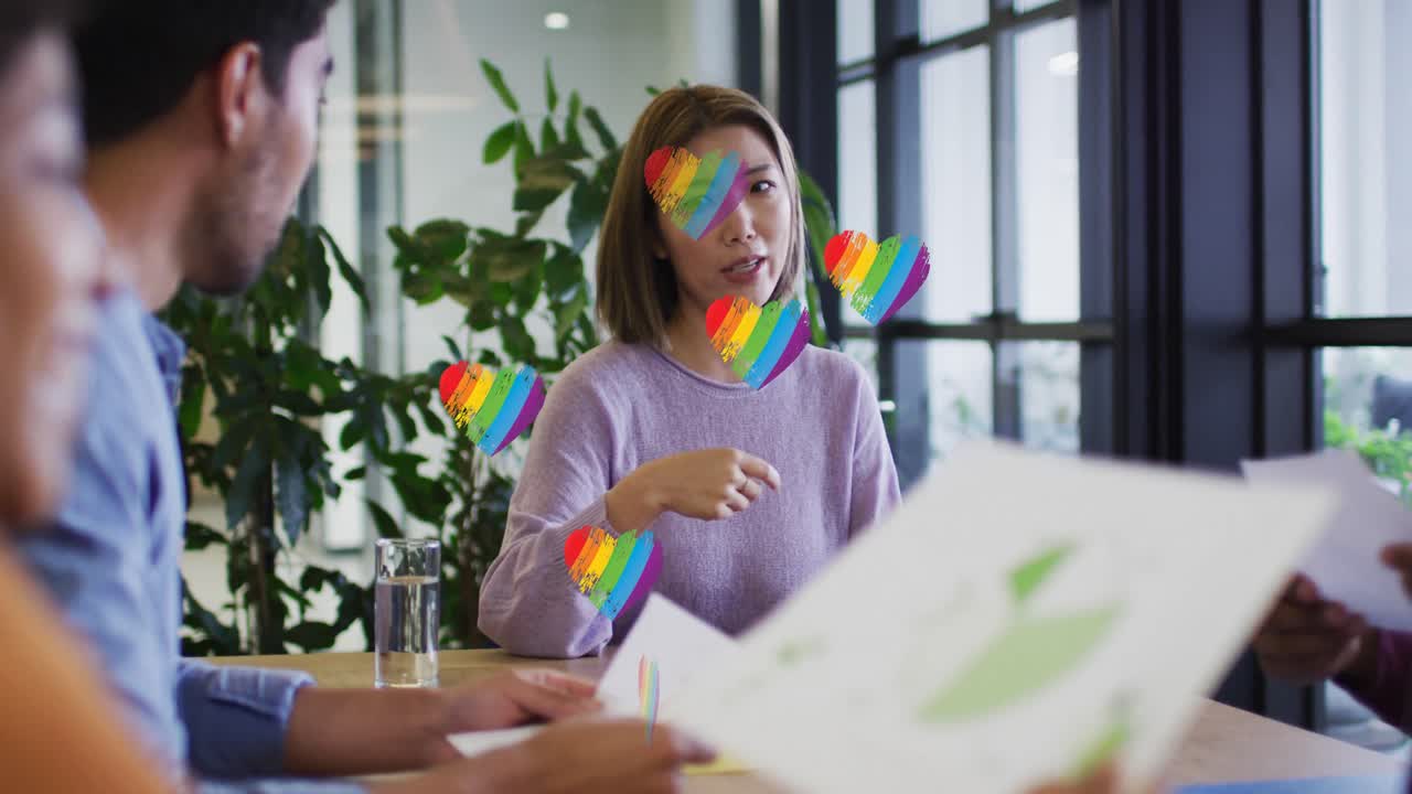 múltiples iconos de corazones arco iris flotando sobre un grupo de diversos colegas de oficina discutiendo en la oficina