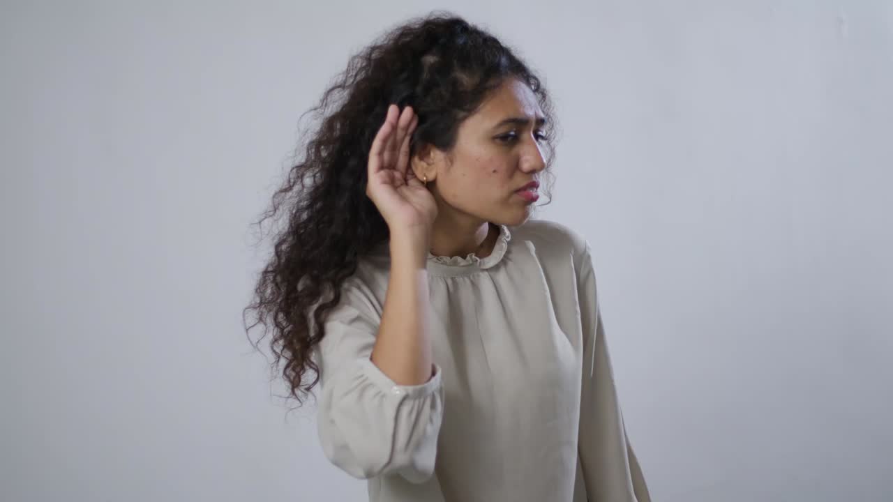 A South Asian woman with curly hair in beige raises her hand and tilts forward as if trying to hear something clearly
