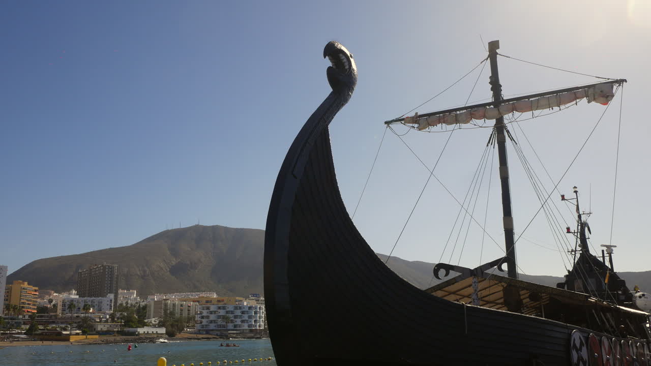Viking ship silhouette forefronts a sunny seaside town, invoking historical nordic boat exploration against a modern backdrop of hotels and mountains in Tenerife