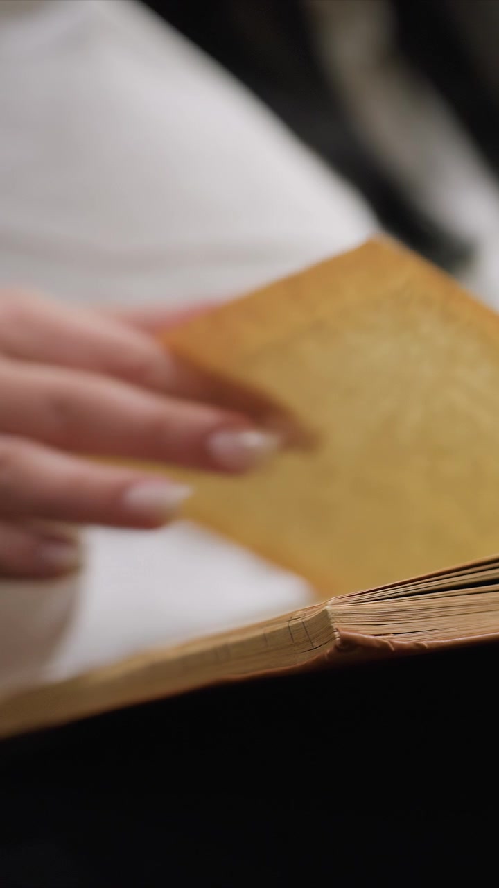 Detail rukou se světlými nehty otáčejících stránky. Tlustý hnědý svazek má mírně opotřebované okraje a zažloutlý papír. Bílé látkové pozadí naznačuje měkké vnitřní prostředí.