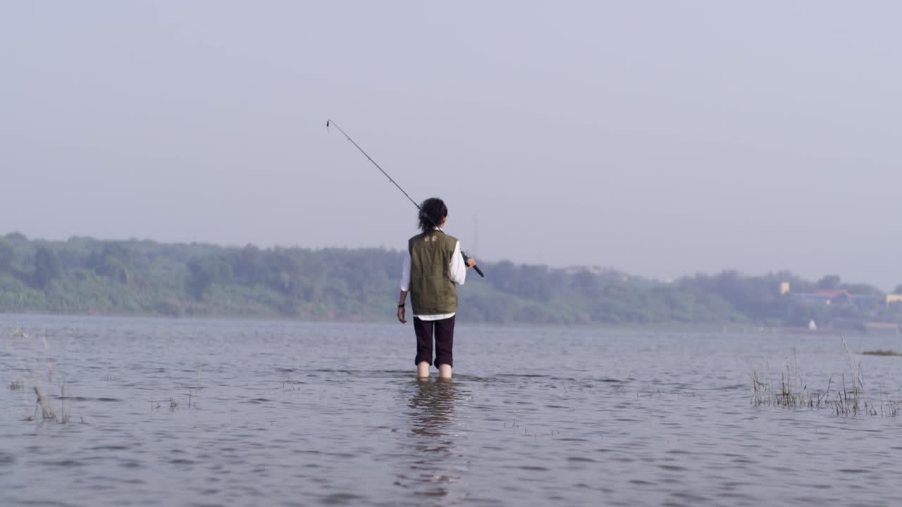 Seorang gadis Asia Selatan mengenakan pakaian kasual luar ruangan berdiri dan memancing di tepi danau yang tenang, bersantai dengan kaki di dalam air