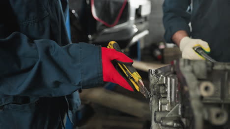 close-up of mechanics in blue uniforms working on an engine, one in red gloves scraping the edge while the other in white gloves hands over a tool and tightens a bolt in an automotive workshop