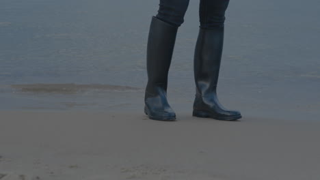 person with gumboot standing on sandy beach with reaching water of baltic sea in background - slow motion low angle shot