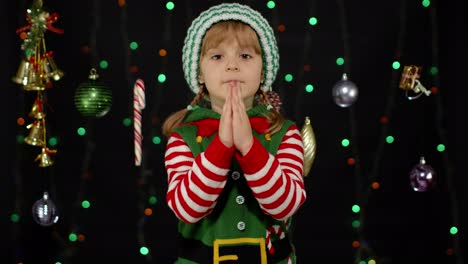 kid in christmas elf santa helper costume begs, prays about gifts on new year. keeps palms together