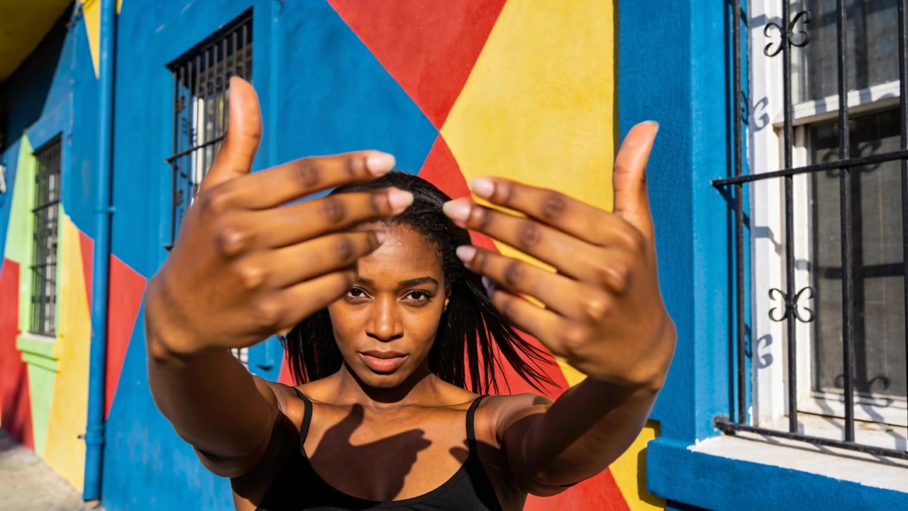 Donna che inquadra e reagisce alla fotocamera, muovendo le mani per incorniciare il viso su un murale sul marciapiede, con canotta nera