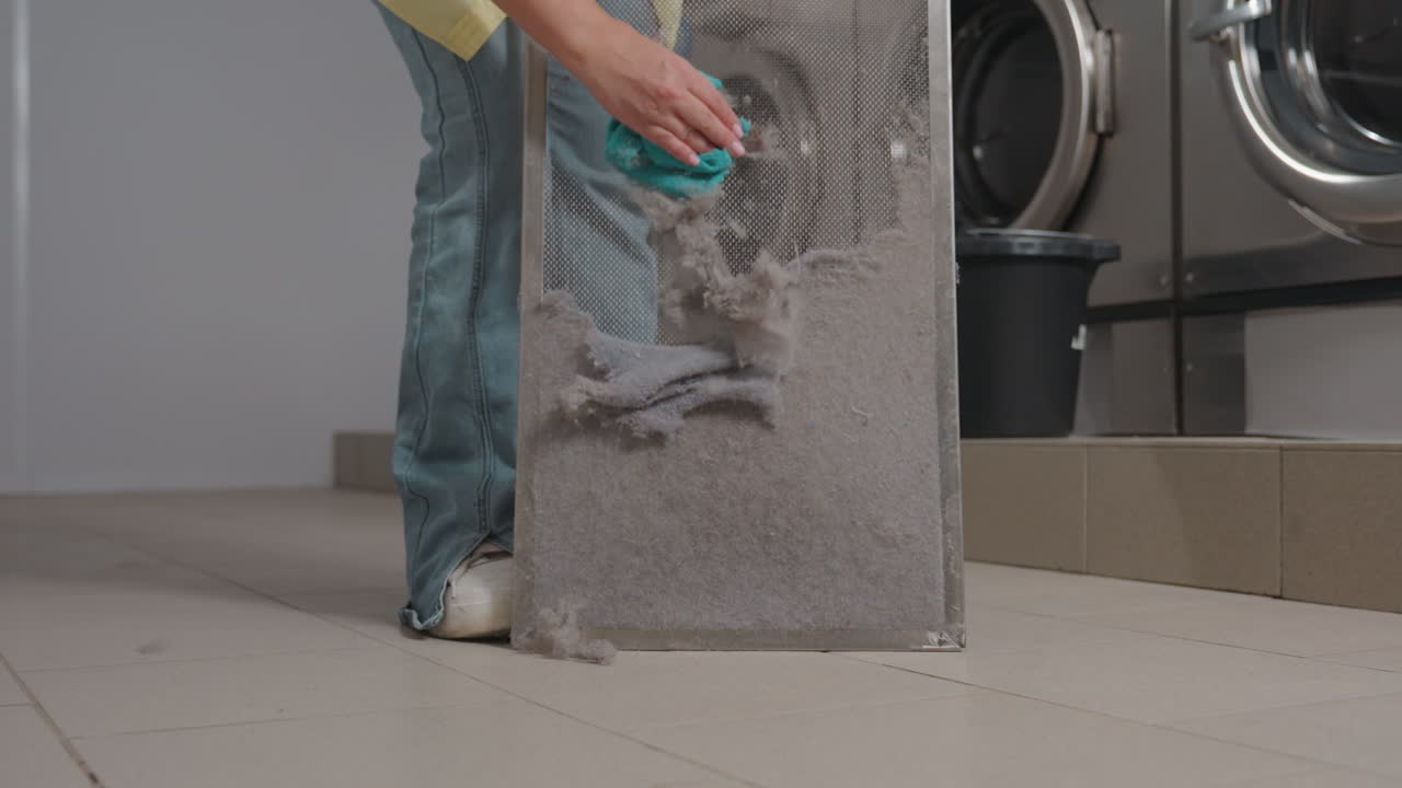 Staff bends to clean washer filter with hand towel, wiping lint dust from mesh, performing maintenance inside laundromat, machines in background, hygiene routine, safety focus, service ethic