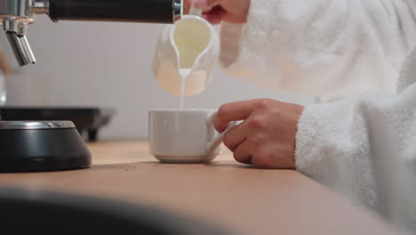 lady pours cream into cup with coffee. woman in terry robe adds milky foam into porcelain cup from jug enjoying early morning. perfect start of weekend