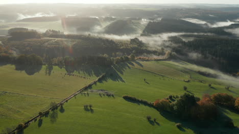 foggy countryside landscape with green fields and forests in sommerain, belgium - aerial pullback