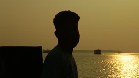 an indian man leans against the wall beside the lake ,sunset time , houseboats in the background , silhouette