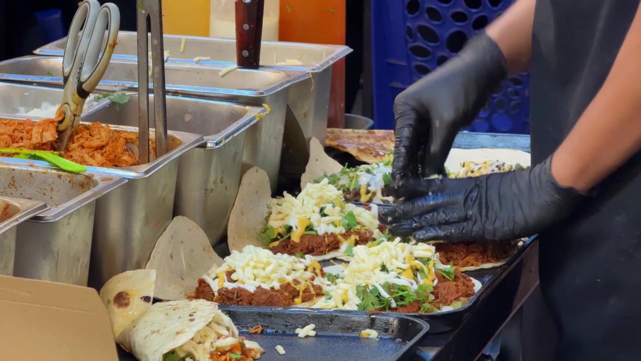 Filmato ravvicinato di un venditore ambulante che prepara numerosi tacos e wrap freschi su una piastra calda. Le tortillas sono riempite con carne, formaggio, verdure, erbe aromatiche e salse durante la frenetica preparazione di cibo da strada.