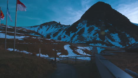 Un-Grupo-De-Banderas-Ondeando-Suavemente-En-El-Viento-En-La-Colina-En-La-Cima-De-Las-Montañas-Grossglockner-Al-Atardecer-En-Austria,-Vista-Amplia