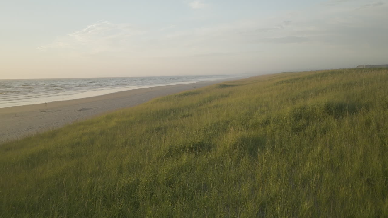 Aerial dolly along sloping edge of grassland plains bordering sandy beach dunes crossing over walking access path