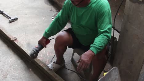wideshot of an old man with white hair cutting a metal bar using an angle grinder with no protective guards like face shield, gloves, shoes and working suit.