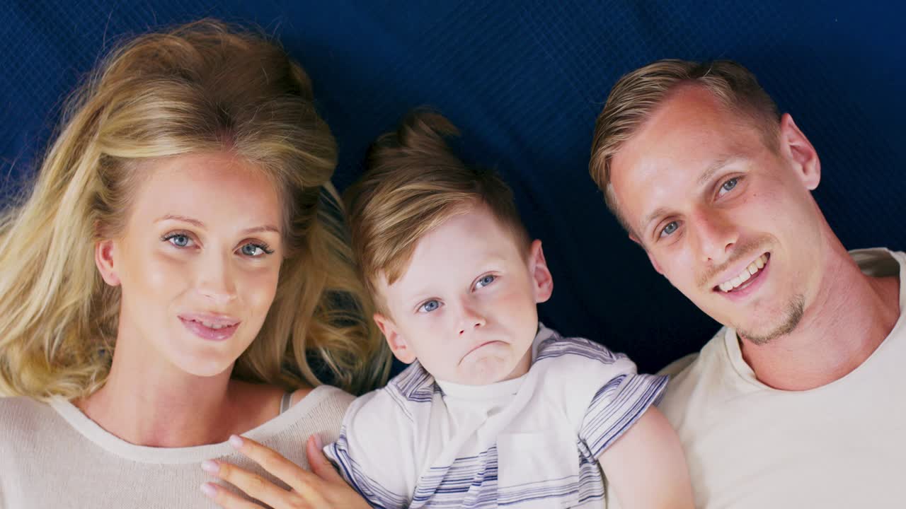 바닥에 누워있는 세 명의 행복한 백인 가족, 부모와 미소 짓는 얼굴의 귀여운 어린 아이가 침실에서 카메라를보고 있습니다.