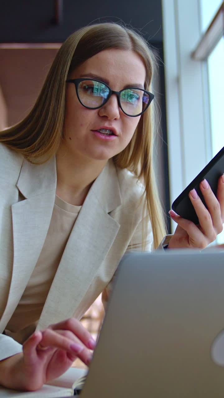 Blonde young woman speaking on the phone leaning on the table. Female employee talks on the phone and works on laptop. Office at backdrop. Vertical video