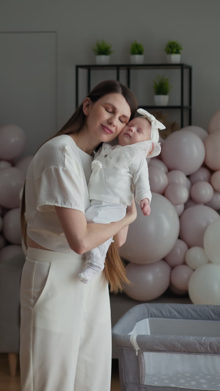 Madre abraza con ternura a su hija bebé dormida, rodeada de suaves globos rosas y blancos