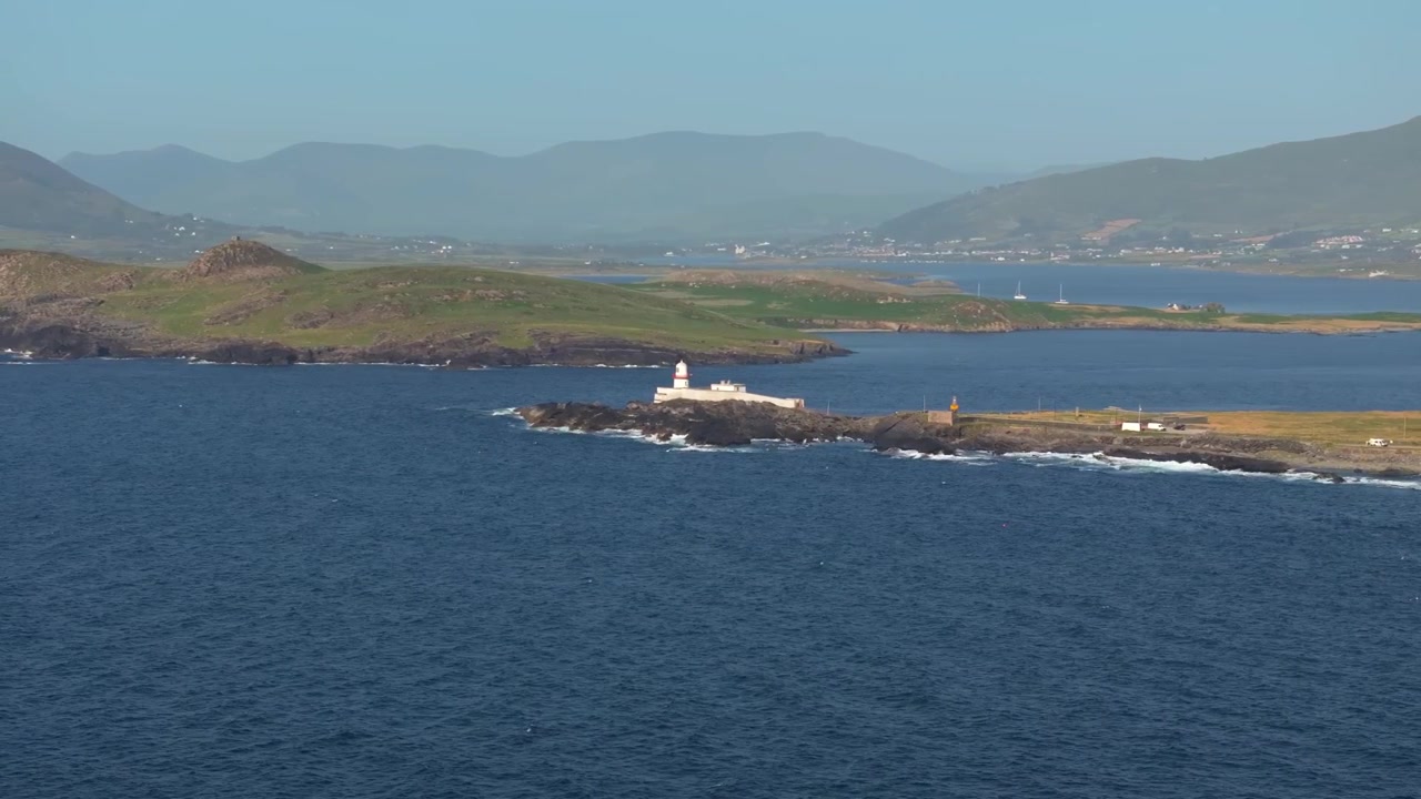 Faro bianco si erge su una punta rocciosa di fronte a un ampio canale di mare blu con colline e isole distanti nella luce soffusa del giorno