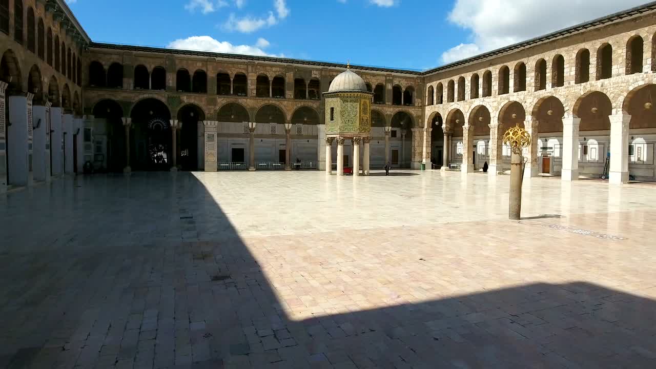 una vista del interior del patio de la mezquita de los umayyad en siria. un avión no tripulado está volando en el patio interior de la mesquita, donde vemos el exterior del edificio dentro de la mezquina.