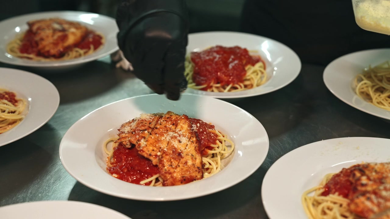 Carrellata media di uno chef con guanti neri che sistema pollo grigliato a fette su spaghetti al sugo di pomodoro marinara e spolvera parmigiano grattugiato sopra.
