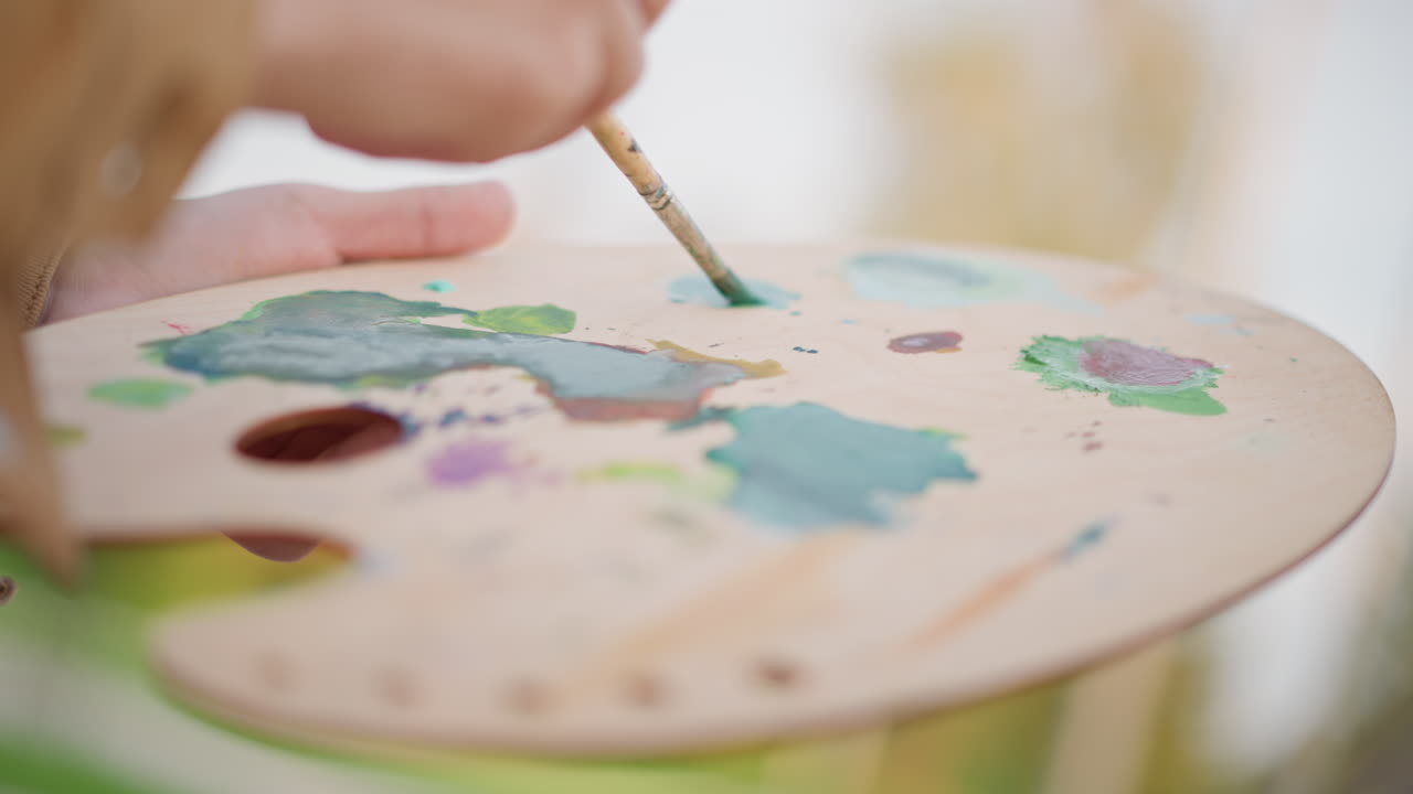close-up de uma paleta com tintas misturadas, artista molhando o pincel em pigmentos verdes e azuis, misturando texturas e pinceladas em um cavalete iluminado pelo sol, poças de pigmentos vívidos, processo criativo capturado em bokeh suave