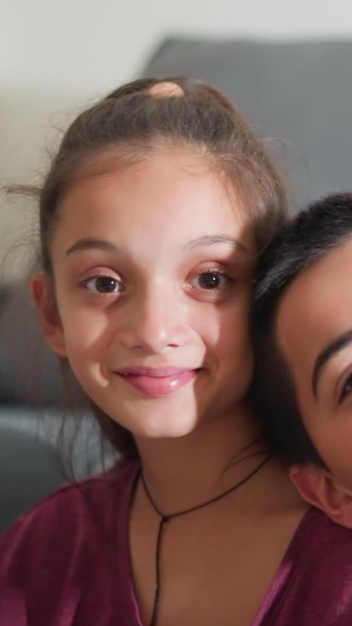 Un hermano pequeño apoya la cabeza en el hombro de su hermana jugando mientras ambos miran a la cámara con una sonrisa dulce, sentados en el suelo contra un sofá gris, mostrando un vínculo fraternal afectuoso.